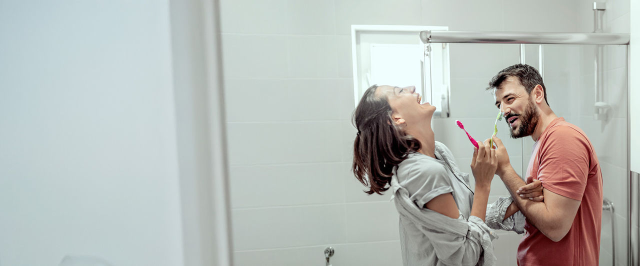 Couple riant dans une salle de bain moderne en se brossant les dents avec des brosses à dents rose et verte.