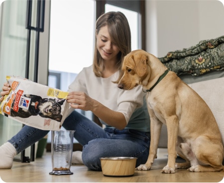 Une femme verse de la nourriture Hill's Pet Nutrition dans un récipient doseur pendant qu'un chien regarde attentivement.