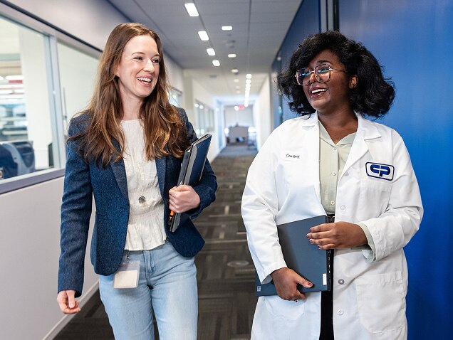 Twee vrouwen, één in vrijetijdskleding en één in een witte laboratoriumjas, lopen en praten in een gang van een Colgate-Palmolive-gebouw, glimlachend en met notitieboekjes in hun handen.